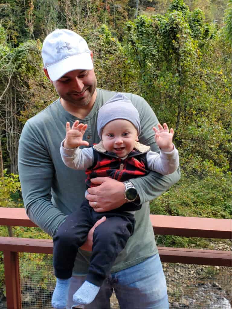 Joey holding Jacob on the balcony overlooking the creek. Jacob is smiling big with both hands in the air.