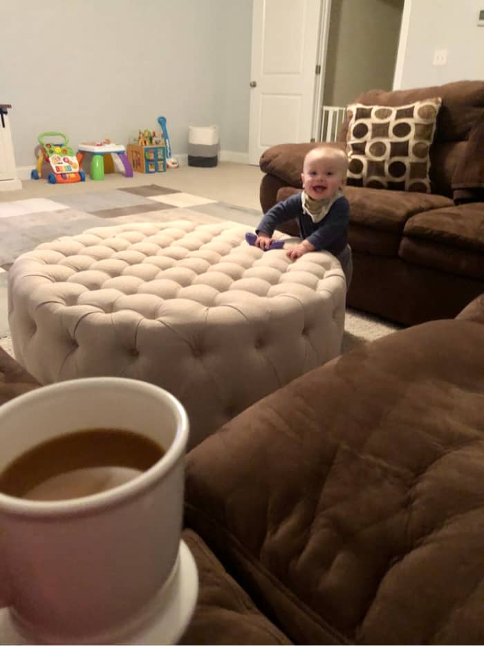 Jacob standing next to the ottoman smiling in the playroom as Lindsey holds her cup of coffee